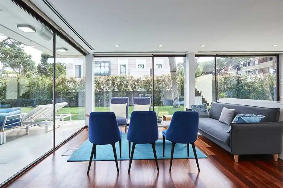 Salón moderno con sillas azules, sofá gris, paredes acristaladas y vistas a un patio y un jardín con tumbonas en el exterior.