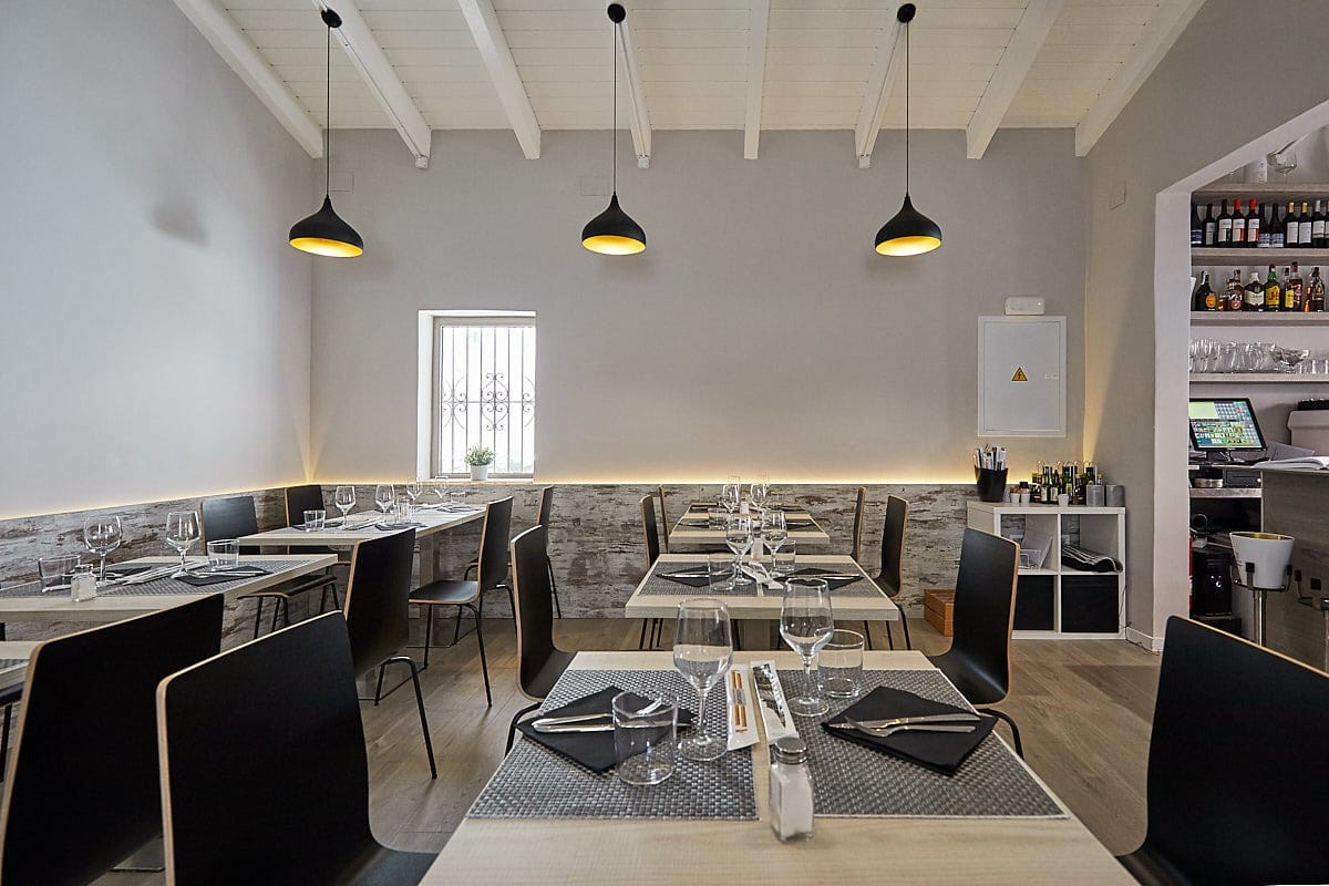 Sala de restaurante con mesas, sillas negras, lámparas colgantes y barra al fondo