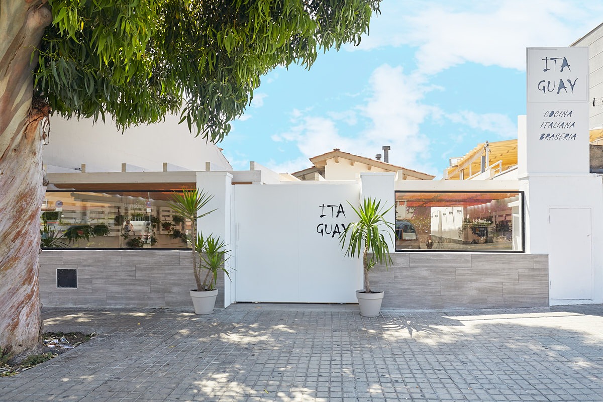 Entrada exterior de restaurante con muro blanco, vegetación y zona de terraza